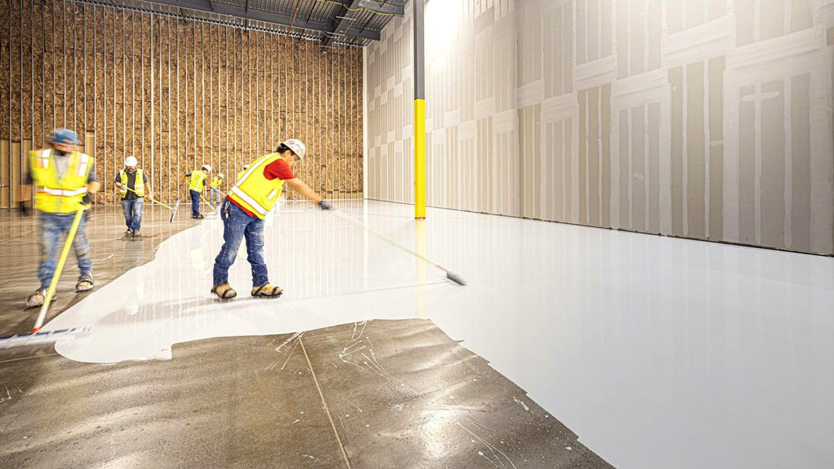 Crew spreading an epoxy flooring coat over a warehouse concrete slab with long-handled rollers